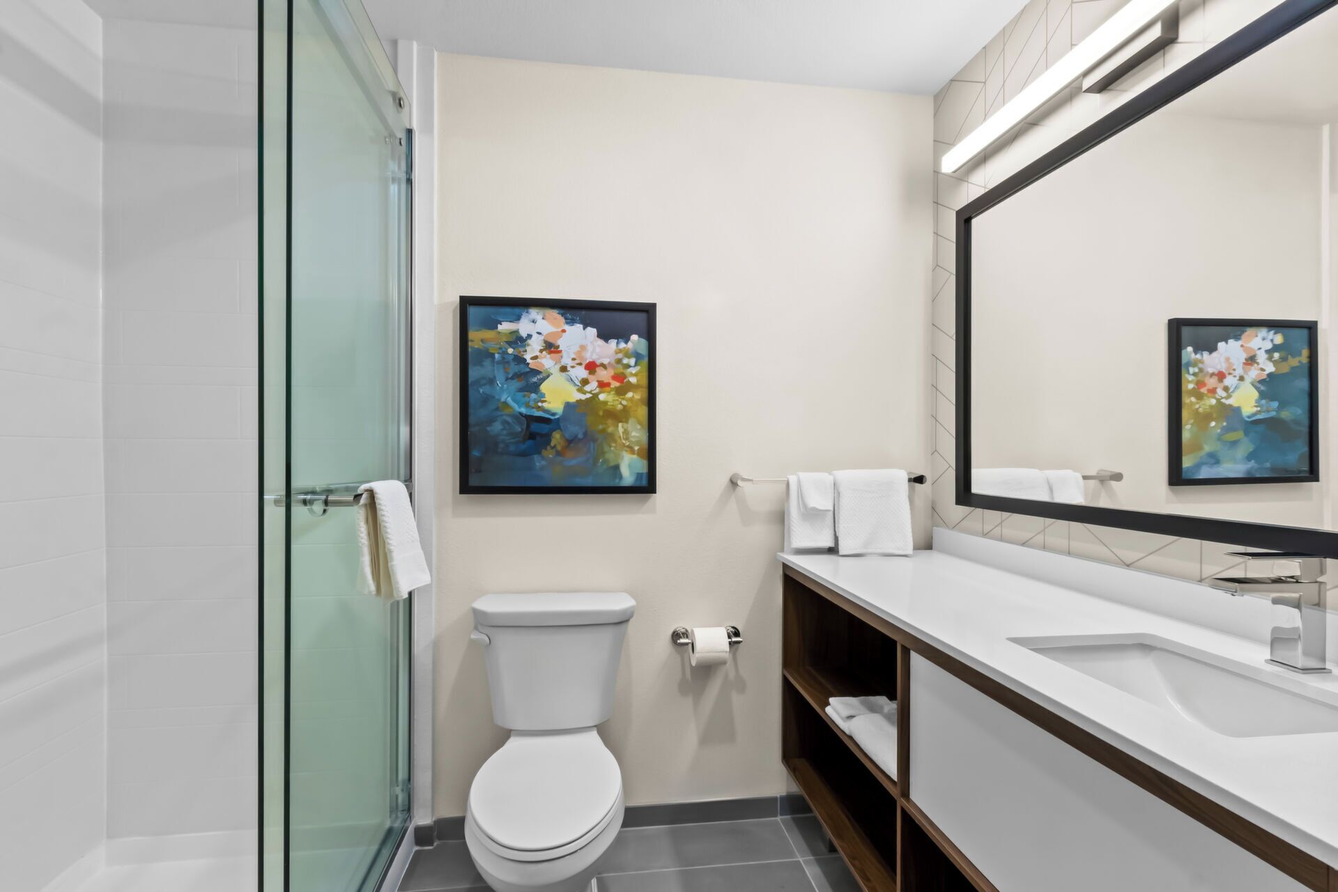 Une salle de bain moderne avec une douche à l’italienne en verre, des toilettes, un lavabo avec un miroir et un tableau abstrait encadré accroché au mur.