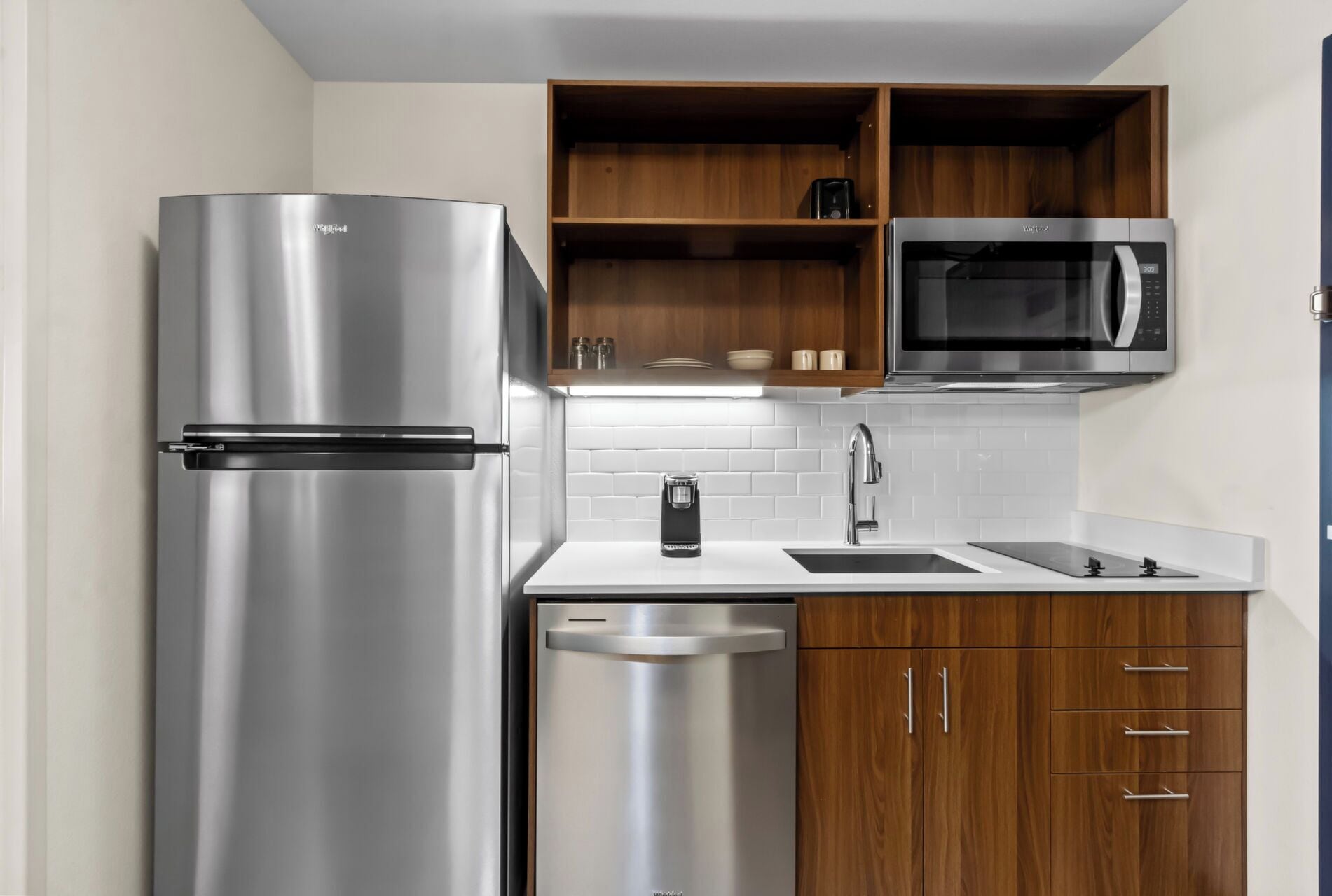 A compact kitchen with a stainless steel refrigerator, a microwave, a coffee maker, and wooden cabinets.