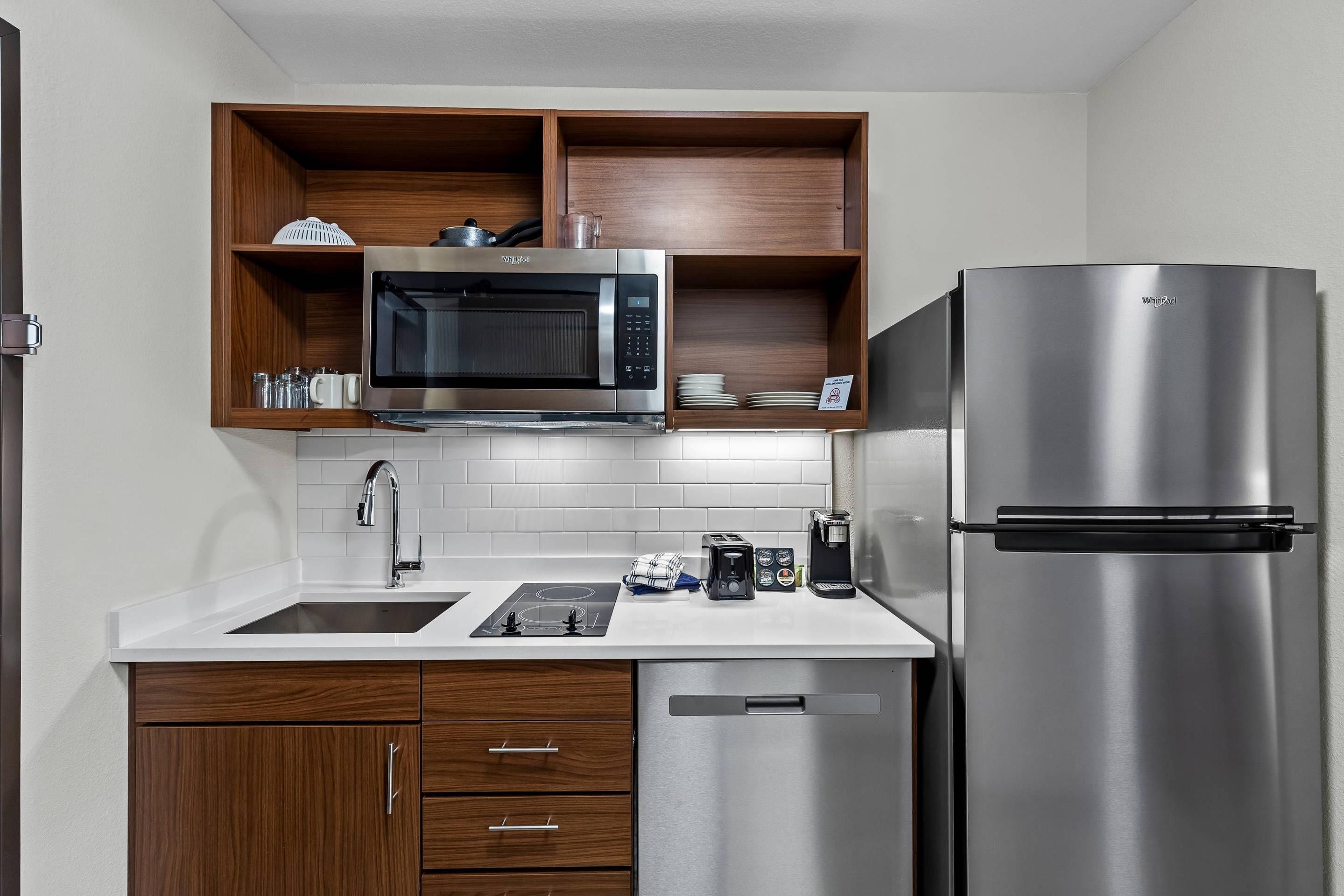 A modern kitchen with a stainless steel refrigerator, a microwave, and a built-in oven. There are wooden cabinets and a white countertop.