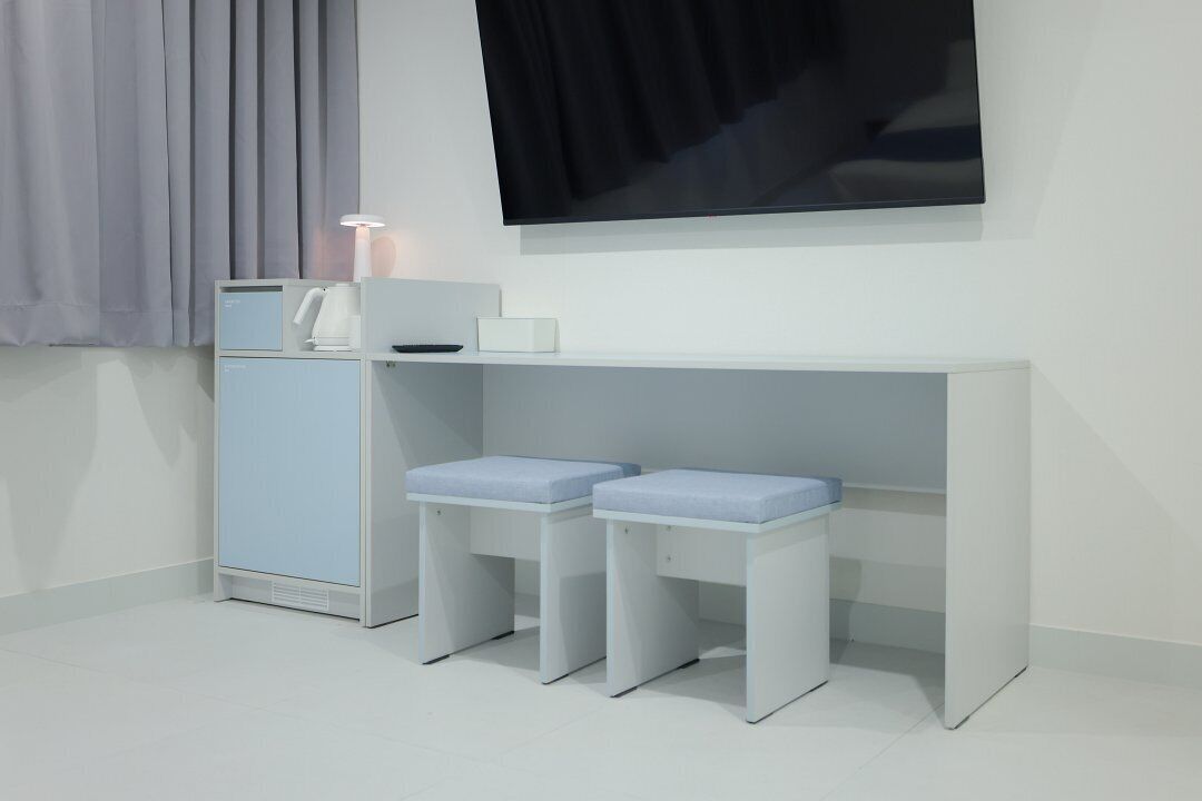 Una habitación minimalista con un escritorio blanco, dos taburetes azules acolchados, un armario blanco y un televisor de pantalla plana montado en la pared.