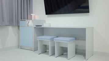 Una habitación minimalista con un escritorio blanco, dos taburetes azules acolchados, un armario blanco y un televisor de pantalla plana montado en la pared.