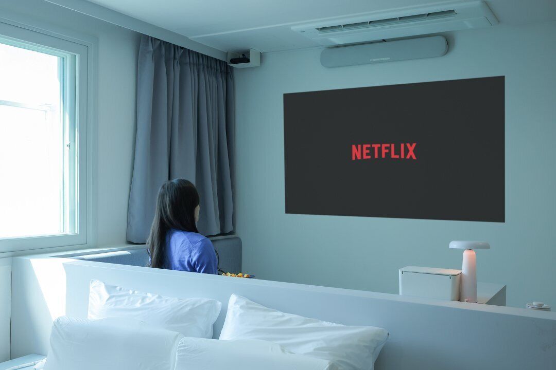 A modern bedroom with a large TV screen displaying "NETFLIX" in red letters, a white bedside table, and a window with curtains.