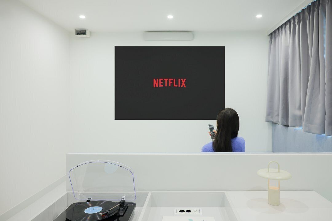 A room with a large screen displaying "NETFLIX" in red letters, a person holding a phone, a turntable, and a small table with a lamp.