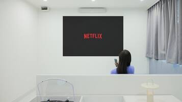 A room with a large screen displaying "NETFLIX" in red letters, a person holding a phone, a turntable, and a small table with a lamp.