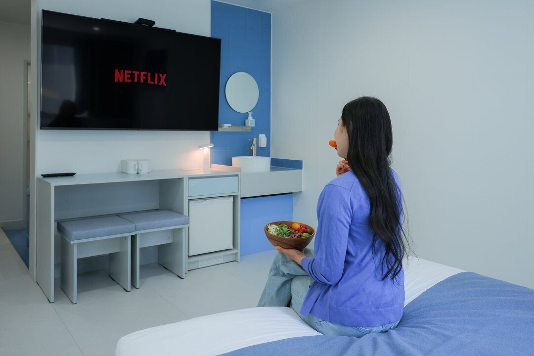 A modern bedroom with a TV displaying 'NETFLIX', a bedside table, a small desk, and a bowl of fruit.