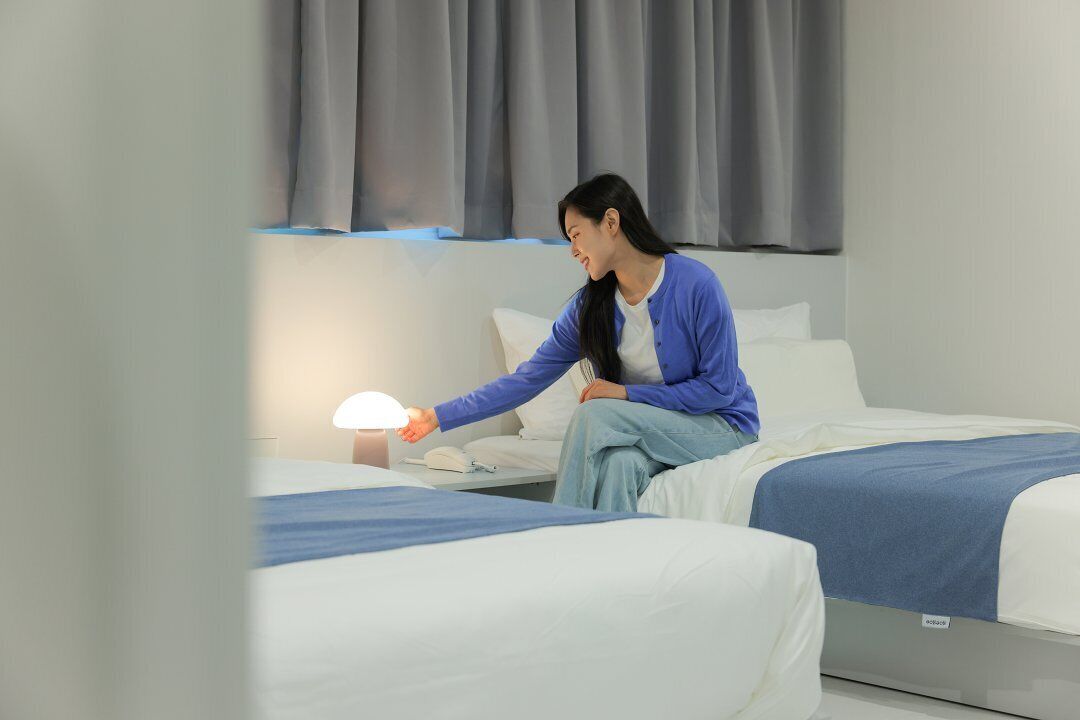 A hotel room with two beds, a bedside table with a lamp, and a window with curtains.