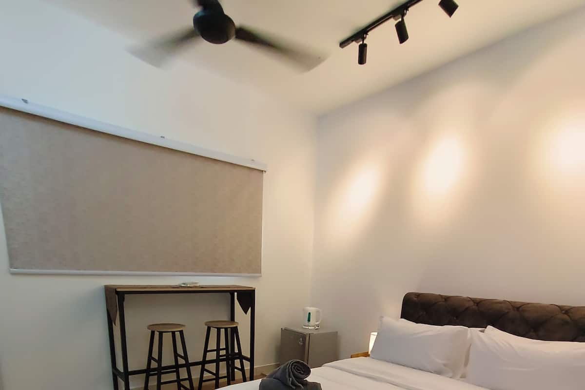 A hotel room with a bed, a desk with two stools, a ceiling fan, and a small cabinet.