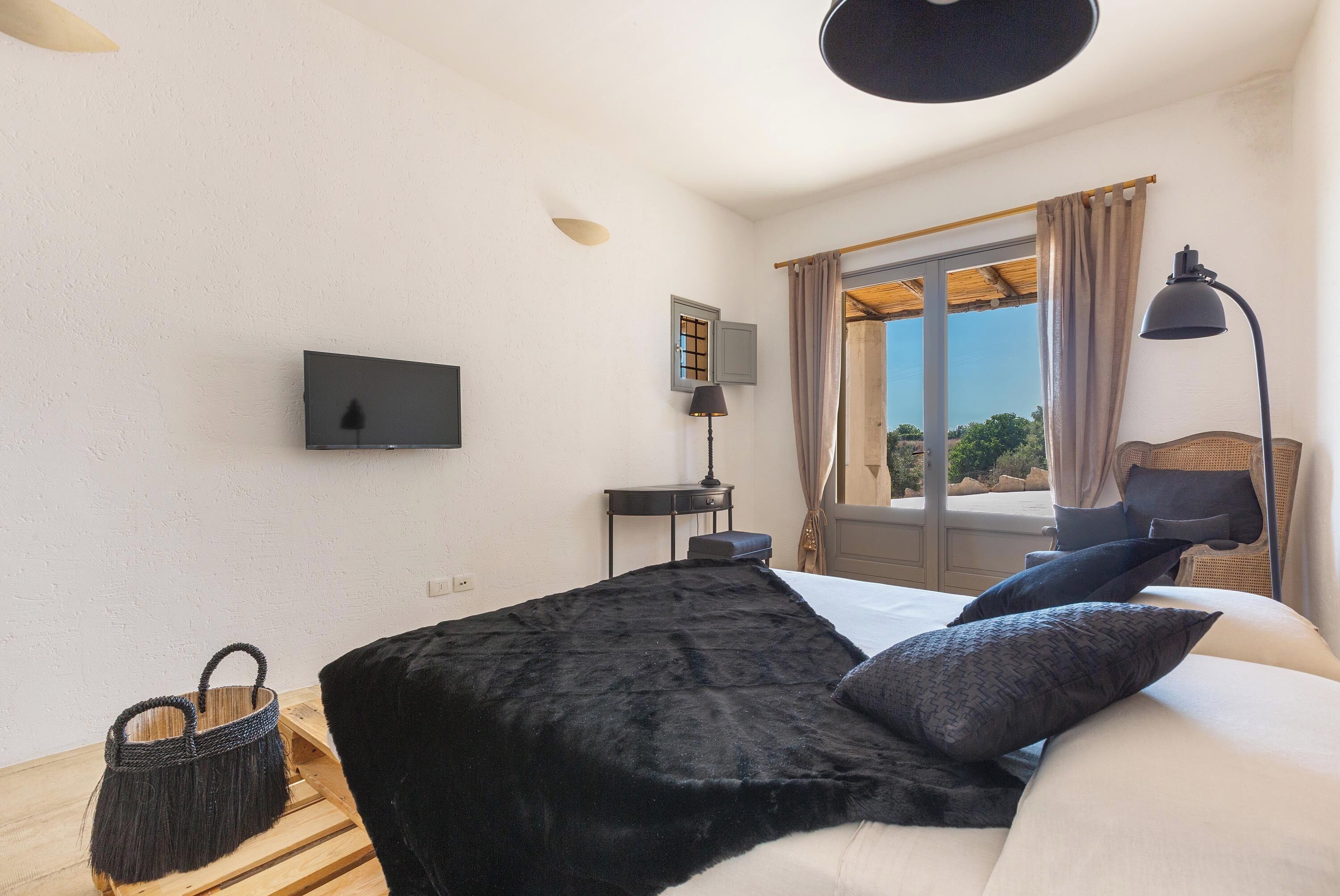 A bedroom with a bed, a television, a small table, a lamp, a window with curtains, and a view of the outdoors.