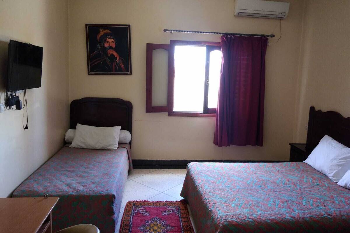 A hotel room with two beds, a TV, an air conditioning unit, and a window with curtains.