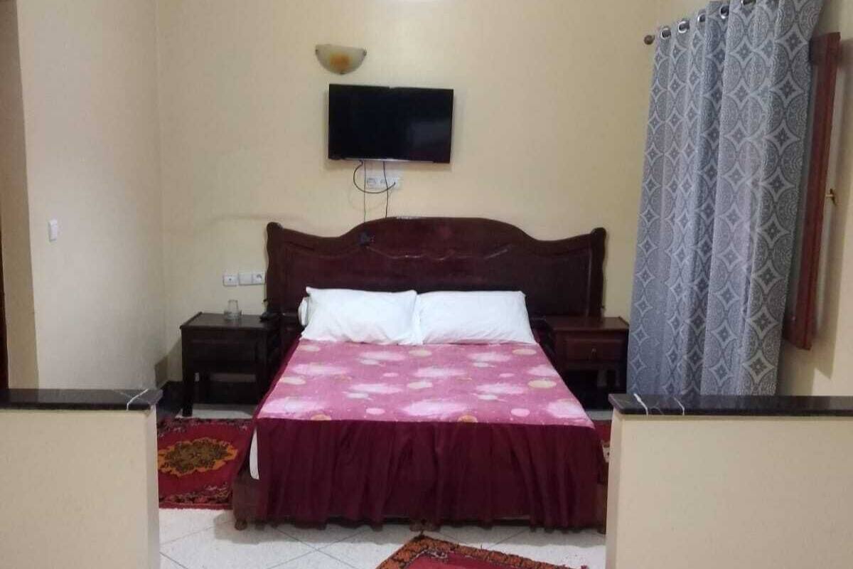 A bedroom with a bed, a television mounted on the wall, a ceiling light, and a patterned curtain.