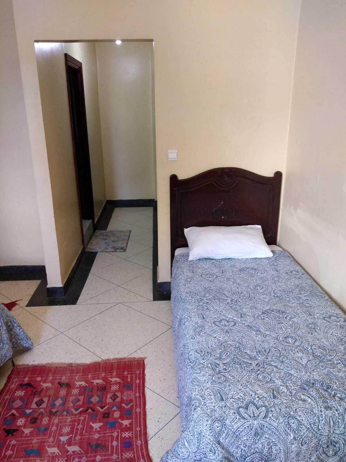 Un dormitorio pequeño y ordenado con una cama individual, un tapete rojo estampado y una pared lisa.