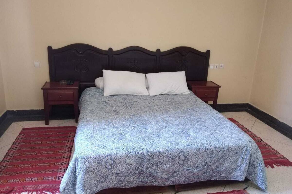 A bedroom with a bed, two nightstands, a red and white patterned rug, and a wall-mounted light fixture.