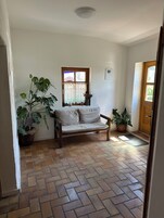A room with a herringbone-patterned floor, a wooden bench with cushions, a window with lace curtains, and potted plants.