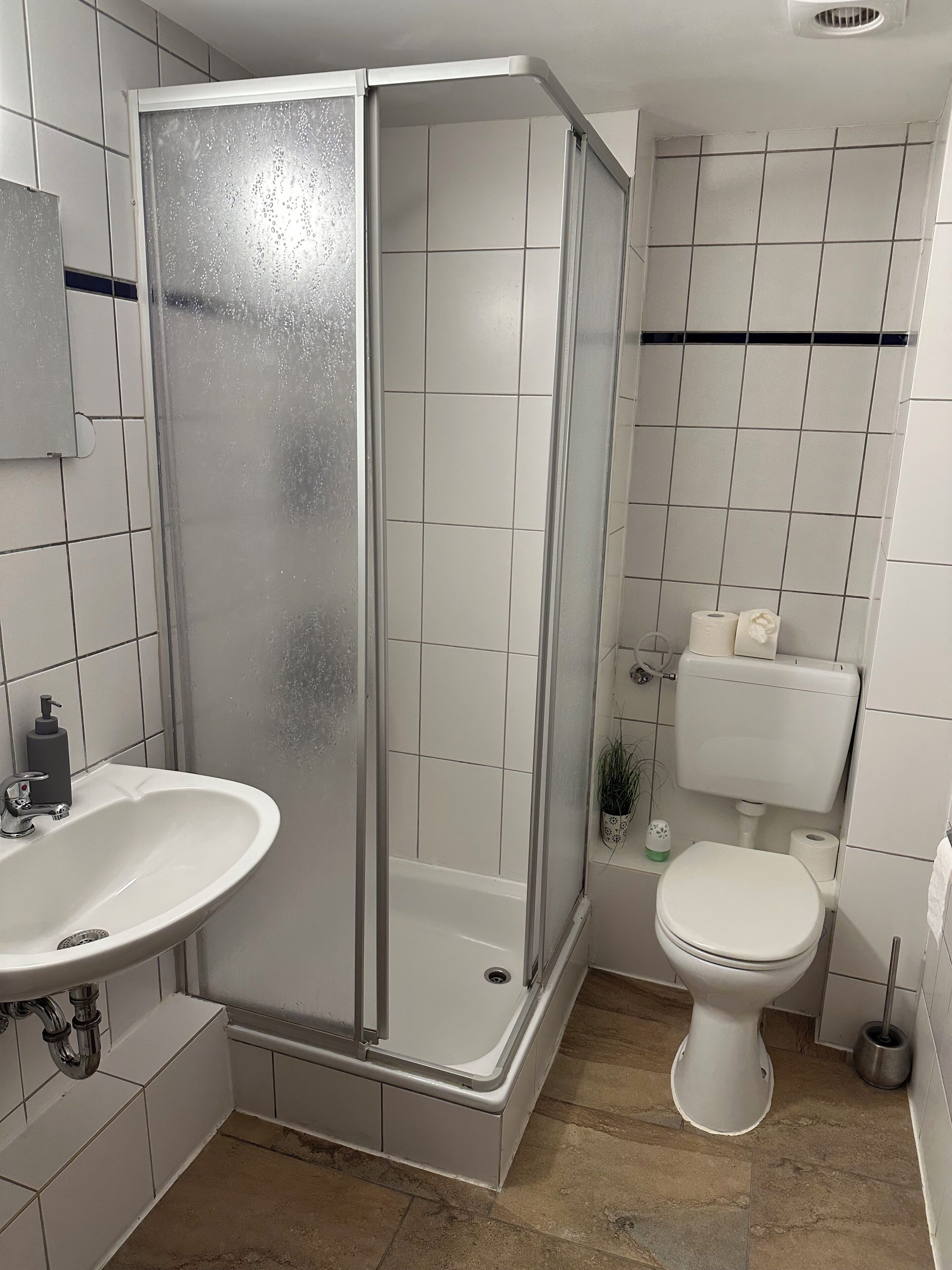 Casa de banho com cabine de duche, lavatório e sanita, incluindo suporte para papel higiénico.