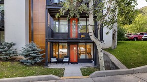 A modern two-story building with a red door, a balcony, and a small outdoor seating area with chairs.