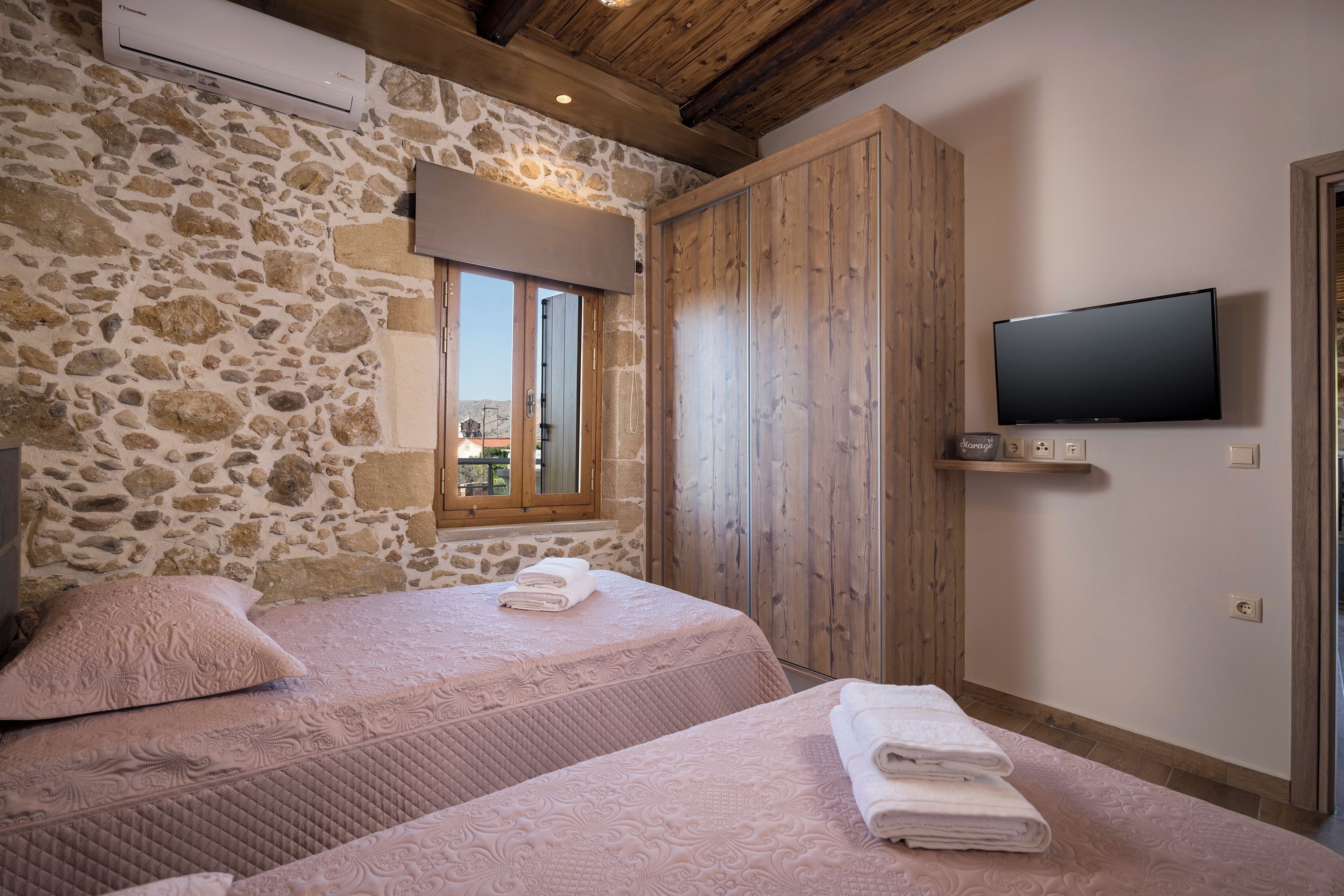 A bedroom with two beds, stone walls, wooden ceiling beams, a TV, and a window with a view.
