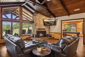 A cozy living room with leather sofas, a stone fireplace, and large windows offering a view of the outdoors. - Serene 3BR Cabin w/ Hot Tub, Game Room, Fishing Pond & 15 Private Acres + Views (Sevierville)