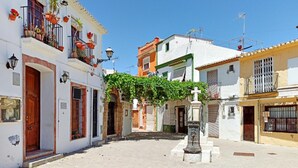 Une rue étroite bordée de bâtiments blancs, de balcons ornés de plantes en pot et d’une statue centrale.