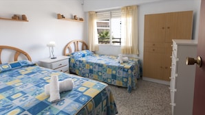 Une chambre avec deux lits, une table de chevet, une commode, une fenêtre avec des rideaux et une vue sur l’extérieur.