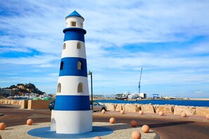 A lighthouse with a blue and white color scheme, situated on a paved area with round bollards, near a body of water and industrial structures. - Llam Ntol in Oliva With 3 Bedrooms and 1 Bathrooms (Oliva)