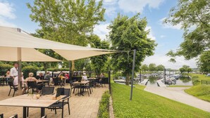 An outdoor patio area with tables and chairs under a large canopy, surrounded by greenery and a body of water. - Hikers Cabin in Maurik (Maurik)