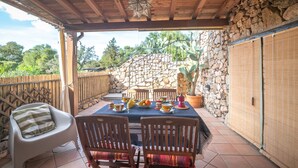 A covered patio with a stone wall, wooden ceiling, and a table set for dining. - Giotto In Sardegna With Shared Pool (Olbia)