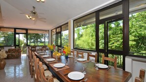 Comedor con una larga mesa de madera dispuesta para seis personas, rodeado de sillas, y grandes ventanas con vistas a zonas verdes.