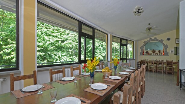Un comedor con una larga mesa de madera dispuesta para una comida, ventanas amplias que ofrecen vista a árboles, y un ventilador de techo.