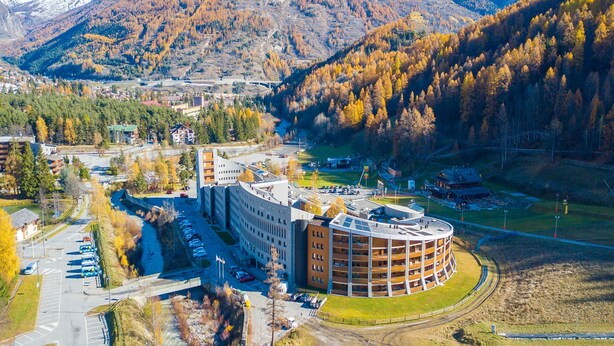 A modern building with a circular central section and a rectangular extension, surrounded by a parking lot and a river. - L Aquila Campo Smith On The Ski Slopes (Bardonecchia)