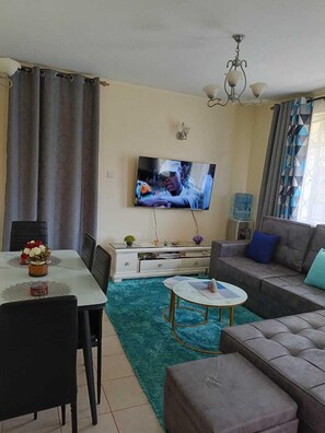 A living room with a TV, sofa, dining table, chairs, and a small coffee table. - Lux Suites MacDade Apartments (Nairobi)