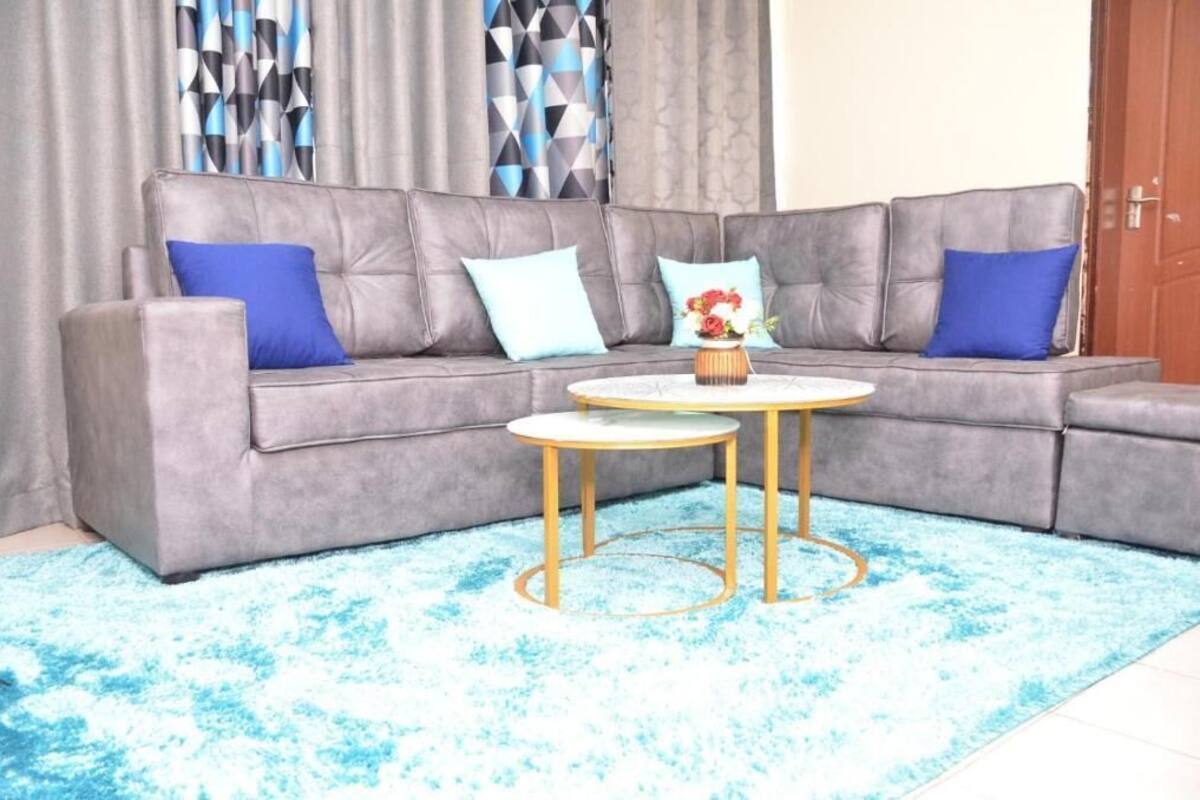 A grey sectional sofa with blue cushions, a small round table with a vase of flowers, and a blue patterned rug.