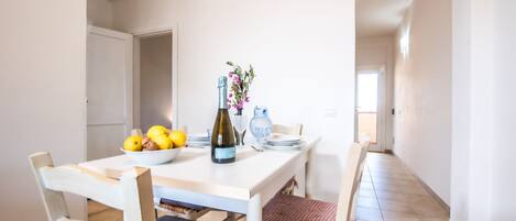A dining area with a table set for two, a bowl of fruit, a bottle of wine, and a vase with flowers.