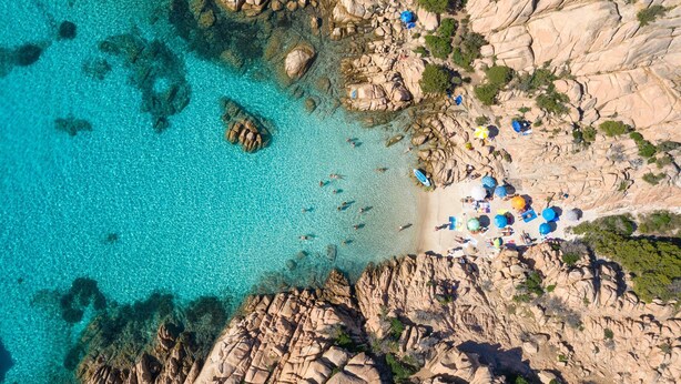 Une vue aérienne d’une plage rocheuse, avec une eau turquoise cristalline, des rochers épars et quelques personnes.