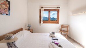 A bedroom with a bed, a window with a view of mountains, a wooden chair, and a painting on the wall. - Monte Majore 16 B in Porto Rotondo (Olbia)