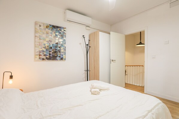 A bedroom with a bed, a wall-mounted air conditioner, a geometric art piece, and a door leading to another room.