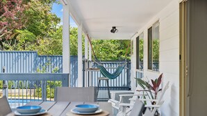 Een overdekte eethoek met witte stoelen en blauwe borden, een hangmat en uitzicht op groen en een blauwe omheining.