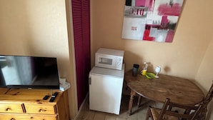 A compact living space with a television on a wooden dresser, a microwave, a small dining table with chairs, and a red door.