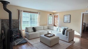 A living room with a fireplace, a sectional sofa, a coffee table, and a large window with blinds. - Maple Cottage at Miller Lake: Your Serene Nature Escape (Miller Lake)