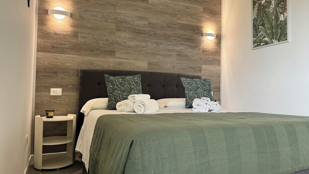 A neatly made bed with a green bedspread, two pillows with a leaf pattern, and a small bedside table with a lamp.