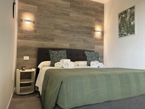 Una cama bien hecha con una colcha verde, dos almohadas con diseño de hojas y una mesita de noche con una lámpara.