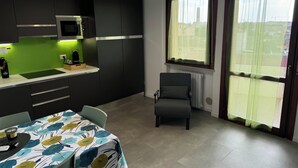 A modern kitchen with dark cabinets, a microwave, and a green backsplash. There is a dining table with a colorful tablecloth and a chair. A radiator is next to a window with a view of the outside.