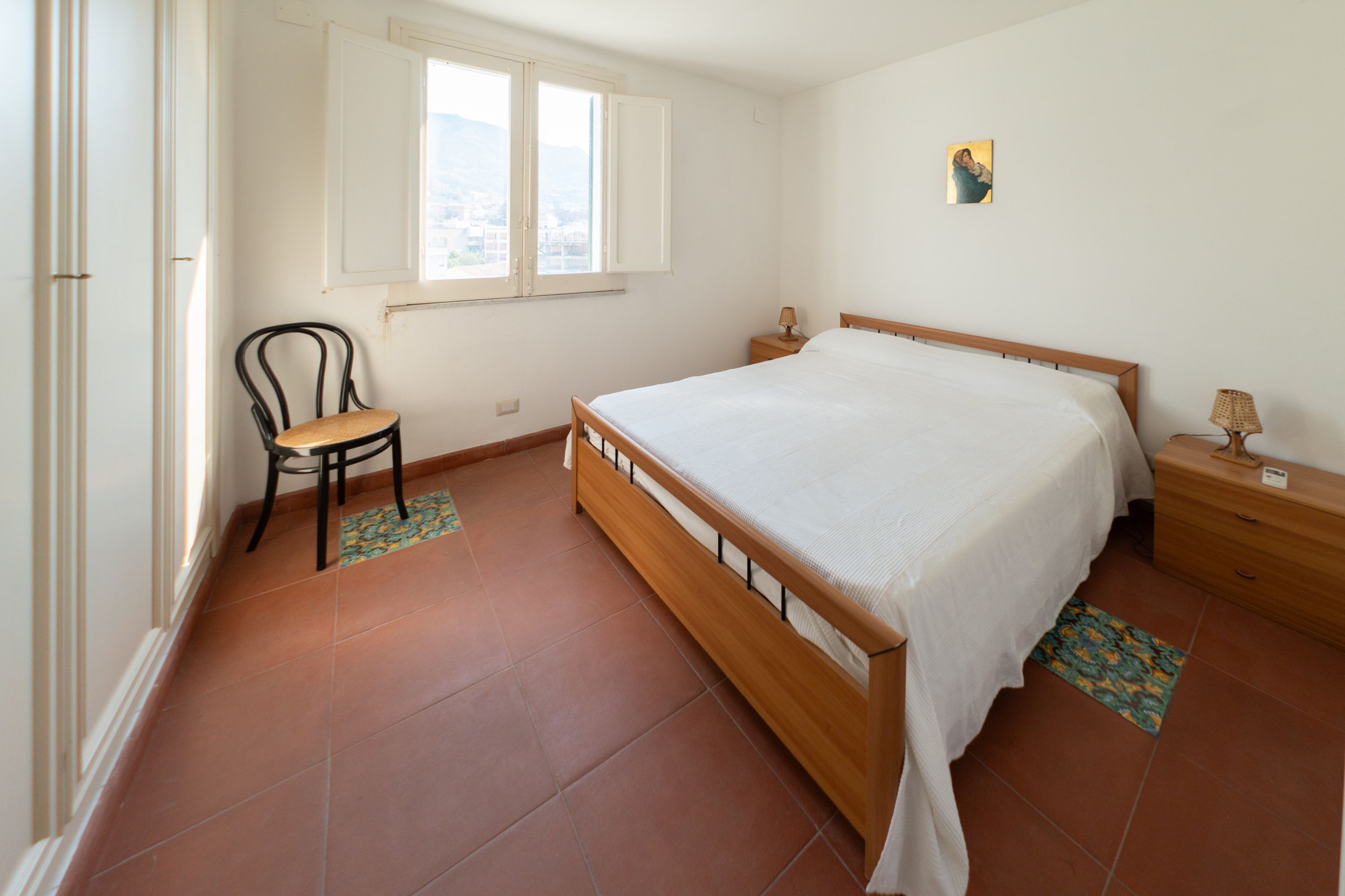 Un dormitorio con una cama de madera, una silla, una mesita de noche, una lámpara y una ventana que ofrece vistas a edificios.