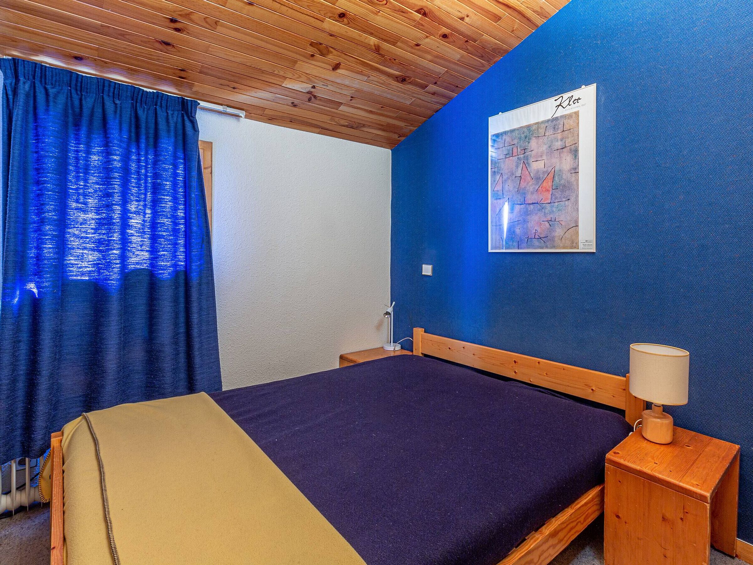 Een kamer met een bed, een nachtkastje, een lamp, een blauwe wand en een houten plafond.