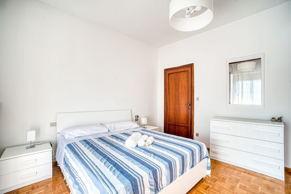 A bedroom with a bed, a wooden door, a white dresser, a nightstand, and a wall-mounted light fixture.