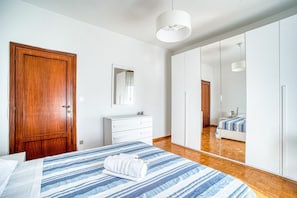 A bedroom with a wooden door, a white dresser, a bed with a striped bedspread, and a large mirror reflecting the room.