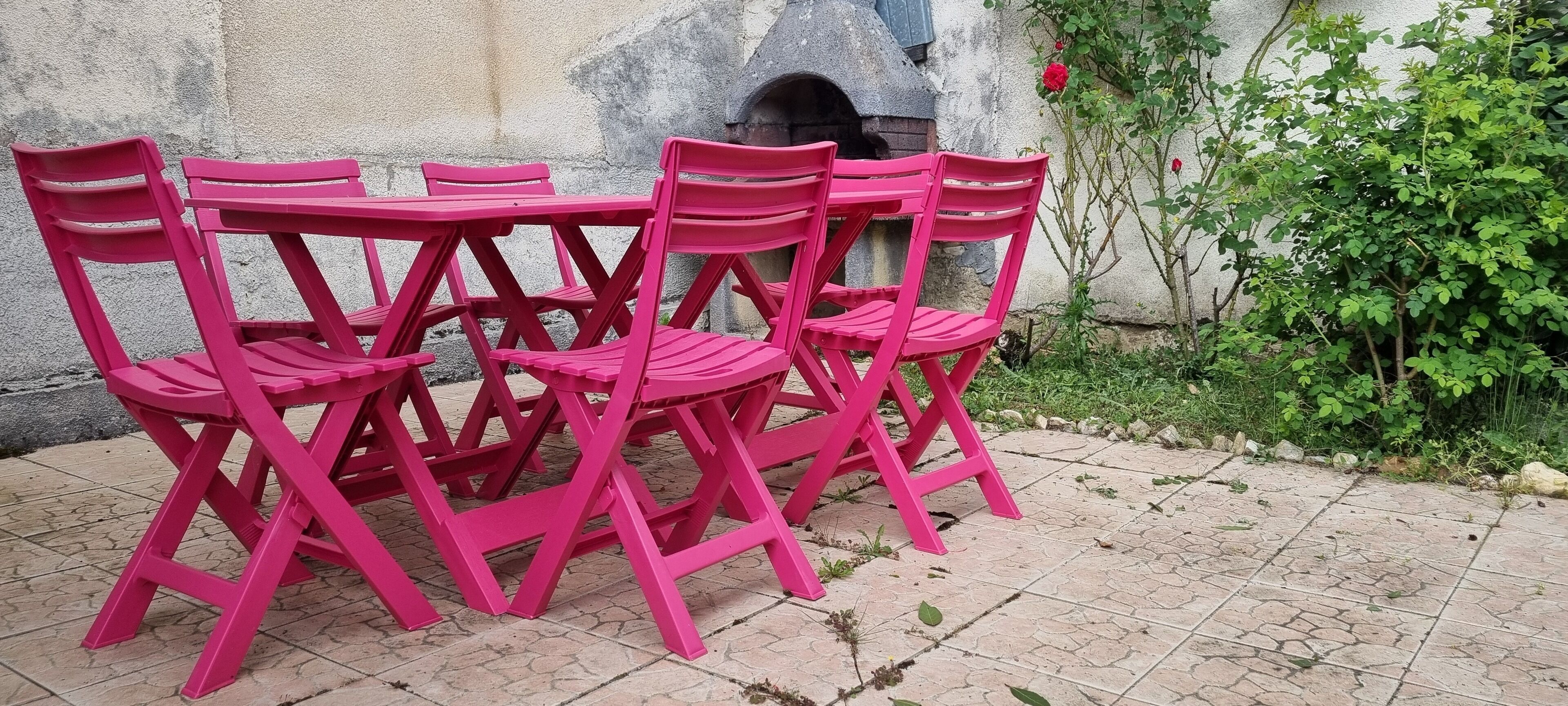 Eine Gruppe aus rosa Stühlen und einem Tisch, draußen auf befestigtem Untergrund platziert, mit einer Steinmauer und Grünpflanzen im Hintergrund.