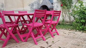 Eine Gruppe aus rosa Stühlen und einem Tisch, draußen auf befestigtem Untergrund platziert, mit einer Steinmauer und Grünpflanzen im Hintergrund.