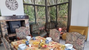 Een eetruimte met een lange tafel, gedekt voor een maaltijd, omringd door gestoffeerde stoelen, een open haard en grote ramen met uitzicht op groen.