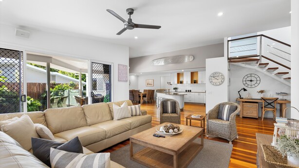 A spacious living area with a sofa, coffee table, and wicker chairs. A kitchen is visible in the background with a dining area.