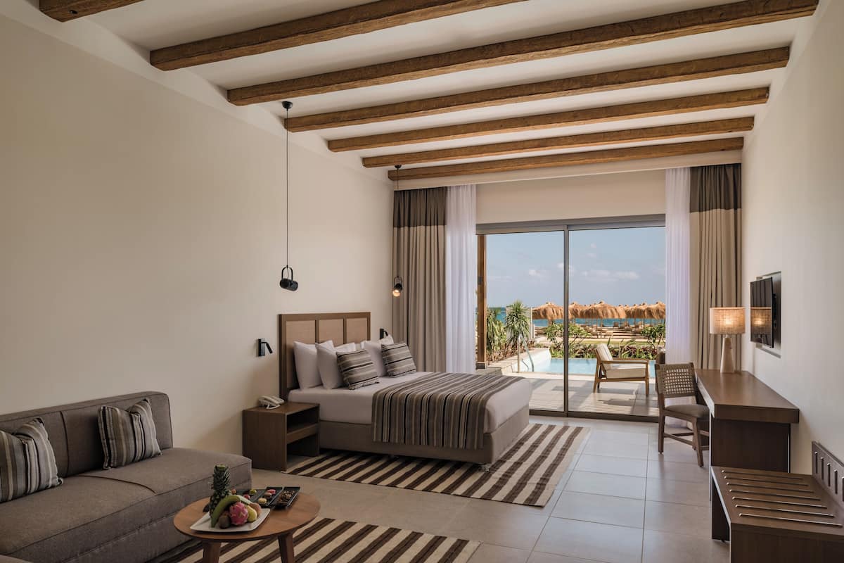 A modern hotel room with a large bed, a sofa, a desk, and a view of a pool and beach.